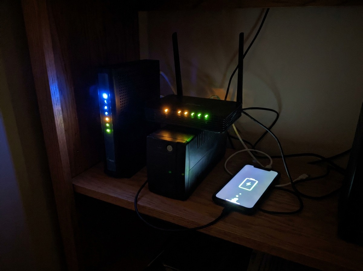 Cable modem and Wi-Fi router on top of a small UPS battery backup with a phone charging beside them during an evening power outage