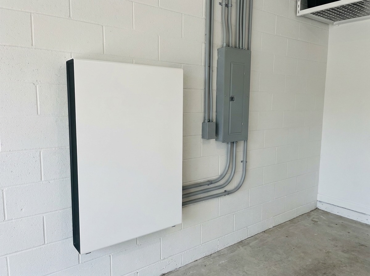 White wall-mounted home battery installed in a garage next to the main electrical panel with no solar panels